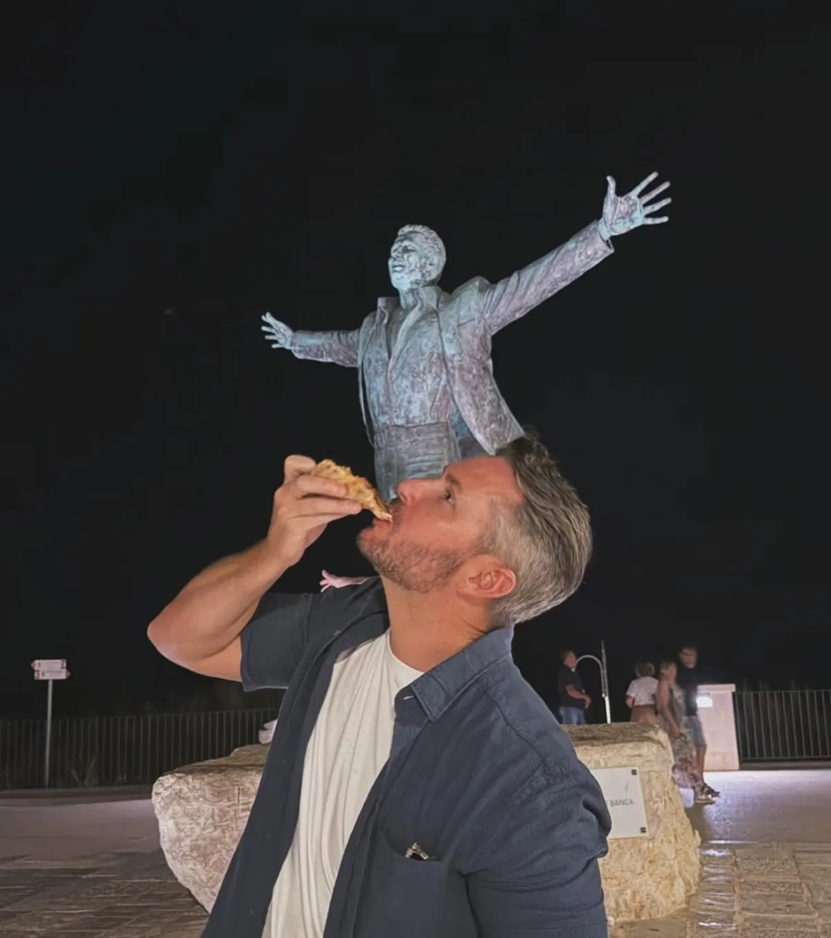 Drew biting into a slice of pizza beneath a statue at night